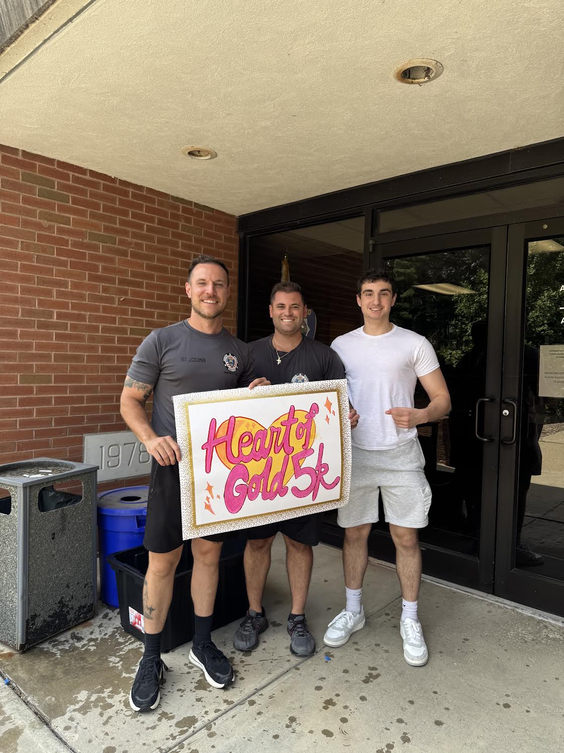 Three people holding a colorful Heart of Gold 5K sign outside a brick building.
